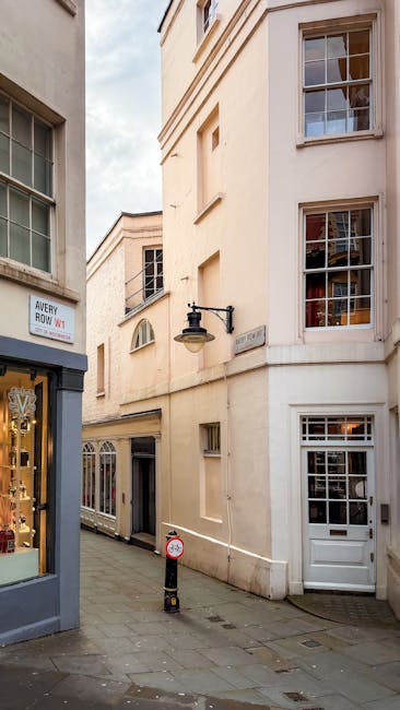 A narrow street scene on Brook Street in Mayfair W1, featuring cream-colored residential buildings with large, multi-pane windows and white wooden frames. The buildings have smooth stucco facades with subtle decorative details and a traditional aesthetic, illuminated by natural daylight. On the left, a shop window displays jewelry, with a sign reading 'Avery How W1' above the entrance. A black wall-mounted lantern is attached to one of the buildings, providing accent lighting. The street surface is paved with stone tiles, and a small bollard with a red and white striped reflective band is positioned near the curb. The overall environment appears clean and well-maintained, reflecting the high standards of domestic cleaning and surface maintenance associated with professional services by Mayfair Cleaners, ensuring hygiene and aesthetic appeal in residential areas.