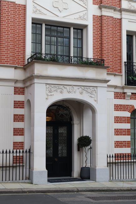 The image depicts the exterior of a historic residential building in Mayfair, W1, featuring a classic brick façade with decorative white stone detailing around the entrance and windows. The black front door is set within an arched white stone surround adorned with intricate carvings, and a small, well-maintained potted topiary sits on the right side of the entrance. Above, a bay window with black framing and patterned glass is visible, and a small balcony with black wrought iron railing extends from the upper floor. The pavement in front is clean, with a black metal fence bordering the property. Natural daylight highlights the building's details, emphasizing the pristine condition of the surfaces, which reflect Mayfair Cleaners' commitment to high standards of cleaning and maintenance for residential properties in the area.