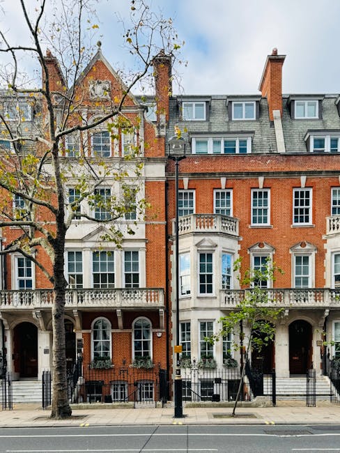 A narrow street scene on Brook Street in Mayfair W1, featuring cream-colored residential buildings with large, multi-pane windows and white wooden frames. The buildings have smooth stucco facades with subtle decorative details and a traditional aesthetic, illuminated by natural daylight. On the left, a shop window displays jewelry, with a sign reading 'Avery How W1' above the entrance. A black wall-mounted lantern is attached to one of the buildings, providing accent lighting. The street surface is paved with stone tiles, and a small bollard with a red and white striped reflective band is positioned near the curb. The overall environment appears clean and well-maintained, reflecting the high standards of domestic cleaning and surface maintenance associated with professional services by Mayfair Cleaners, ensuring hygiene and aesthetic appeal in residential areas.