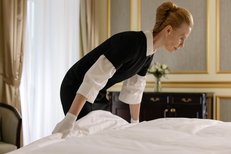 A professional cleaner from Mayfair Cleaners, wearing a black and white uniform and white gloves, is carefully preparing a bed in a well-lit bedroom for Airbnb turnover. The scene shows the cleaner sm