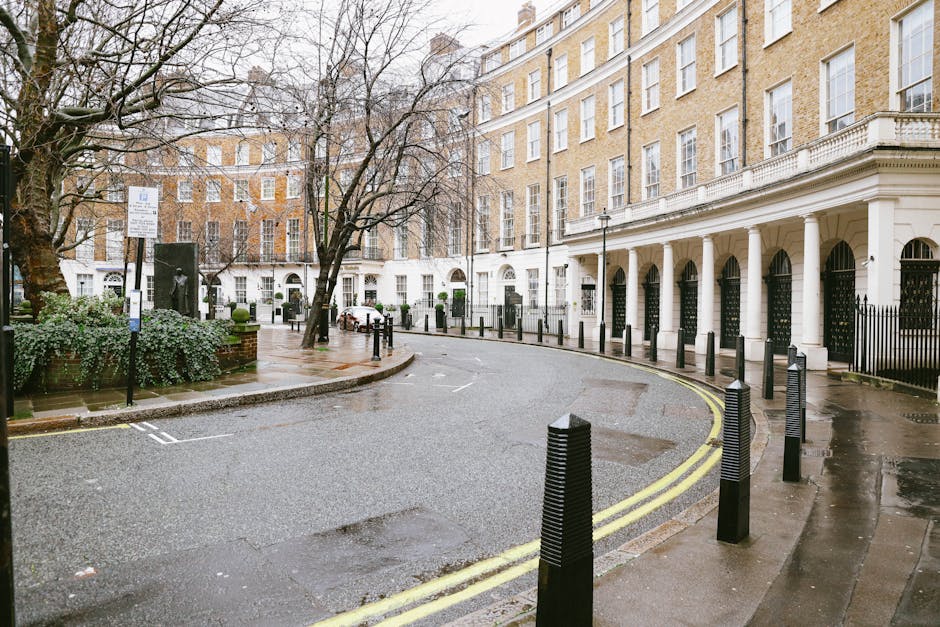 Photograph of a street scene outside residential buildings in Grosvenor Square, Mayfair, featuring red-brick Victorian-style terraced houses with large bay windows, some with decorative white trims. T