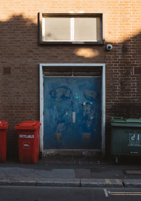 A row of four wheelie bins for waste separation, positioned on a cobbled street in front of an orange building with barred windows. The bins are made of plastic and have different colored lids—white